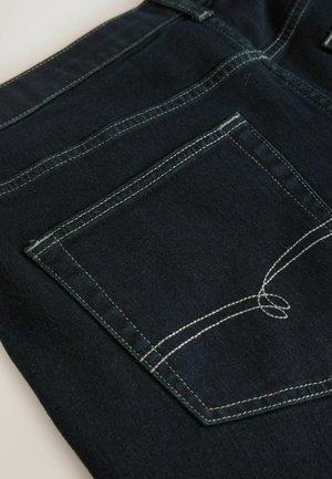 Sügavsinine teksakangas kontrastsete valgete õmblustega. Omab tagakotti, millel on tikandiga detail ja ruudukujuline vorm.