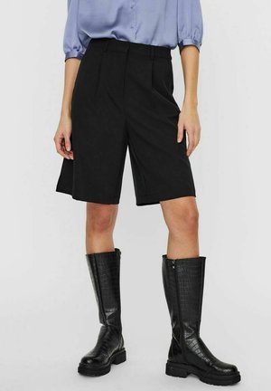 Femme portant un short noir cintré longueur genou, un chemisier bleu clair à manches bouffantes et des bottes noires texturées montantes jusqu’au genou.