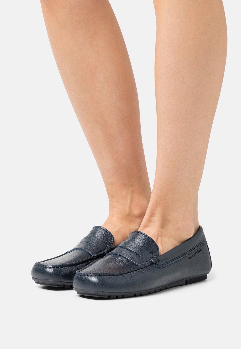 Pieds portant des mocassins en cuir bleu foncé à enfiler avec détails de coutures et semelles basses en caoutchouc sur un fond clair uni.