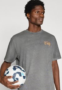 Γκρι βαμβακερό μπλουζάκι με στρογγυλή λαιμόκοψη, με την επιγραφή "City" σε πορτοκαλί γραφή στο στήθος. Το άτομο κρατά μια λευκή και μπλέ μπάλα ποδοσφαίρου.