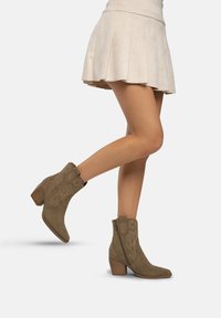 Olivgrüne Wildleder-Stiefeletten mit spitzem Zehenbereich, dekorativen Nähten und einem seitlichen Reißverschluss, kombiniert mit einem plissierten cremefarbenen Rock.
