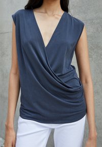 Navy-blaues Oberteil mit tiefem V-Ausschnitt und kurzen Ärmeln, aus weichem Stoff, kombiniert mit weißen, taillierten Hosen. Glatte Textur, subtile Drapierungen.