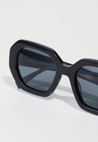 Black octagonal sunglasses with dark lenses. Thick plastic frame features a smooth finish. Brand name displayed on the inner arm.