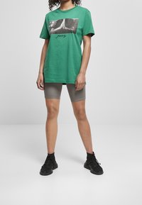 Camiseta de algodón verde con una impresión fotográfica y el texto "reza", combinada con pantalones cortos de ciclismo grises y zapatillas negras.