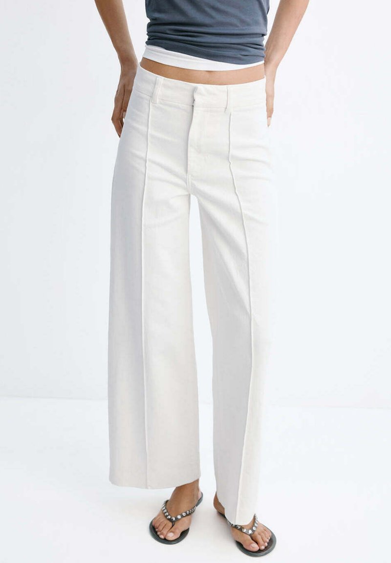 Personne portant un pantalon blanc taille haute à jambes larges avec coutures avant, associé à un top court bleu et des sandales noires avec des clous argentés.