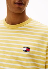 Kollane ja valge triibuline lühikeste varrukatega T-särk ümmarguse kaelusega, millel on väike ruudukujuline logopatch tekstiga: "TOMMY JEANS."