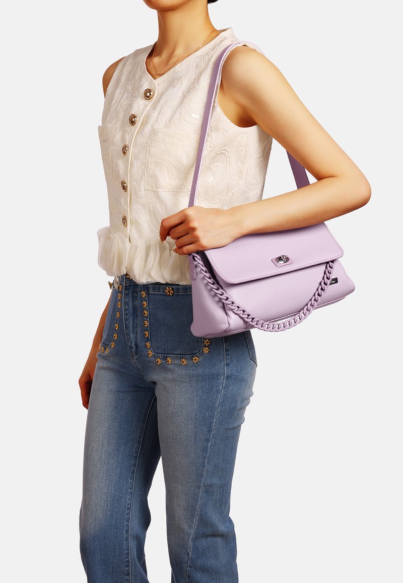 Borsa a mano in pelle lilla con dettaglio a catena intrecciata, chiusura a patta e piccola ferramenta argentata. Indossata con una blusa bianca testurizzata e jeans decorati.