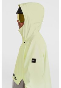 Gul vandtæt jakke med hætte, grå nederste del og sort logo-patch. Har en frontlomme og et slankt design til udendørs brug.