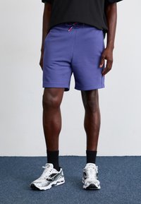 Shorts de sport violets avec cordon de serrage, en tissu léger, texture lisse, accompagnés de chaussettes noires à la cheville et de baskets blanches/argentées.