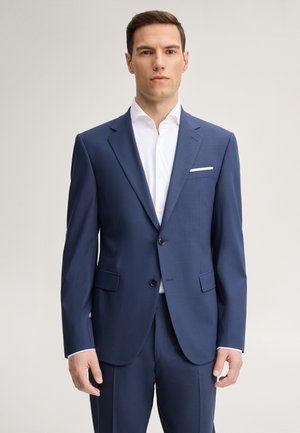 Uomo in piedi rivolto in avanti, indossa un abito blu e una camicia bianca, con un fazzoletto bianco nel taschino, su uno sfondo chiaro e semplice.