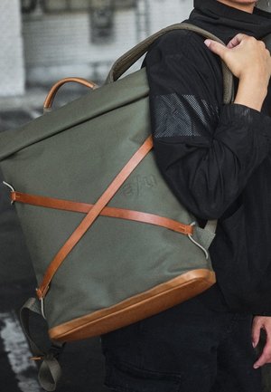 Persona con chaqueta negra que lleva una mochila grande verde oliva con correas y base de cuero marrón claro, sujetando una correa del hombro.