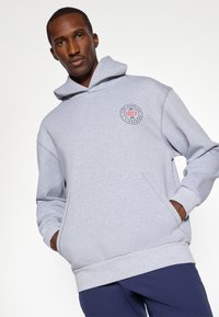 Lys grå hettegenser med en frontlomme, snor i hetten og et sirkulært logo på brystet med røde og blå detaljer.