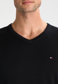 Czarny sweter z dekoltem w kształcie litery V, wykonany z teksturowanej tkaniny, z małym czerwono-białym-niebieskim logo po lewej stronie klatki piersiowej. Prosty i elegancki design.