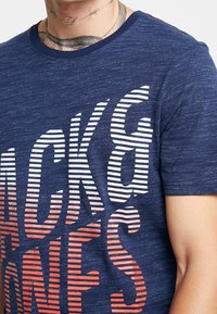 Mu muž nosí námořnickou modrou tričko s kulatým výstřihom, na kterém jsou bílé a červené pruhy textu, částečně viditelné tetování na krku a odkryté rameno.