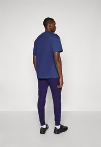 Marineblauw katoenen T-shirt met korte mouwen, gecombineerd met paarse slim-fit joggers. Zwarte sportschoenen met witte sokken maken de look compleet.