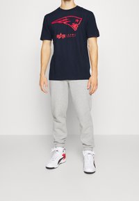 Námořnické tričko s červeným logem a nápisem, šedé tepláky, bílé a červené tenisky. Neformální sportovní oblečení. Krátké rukávy a uvolněný střih.