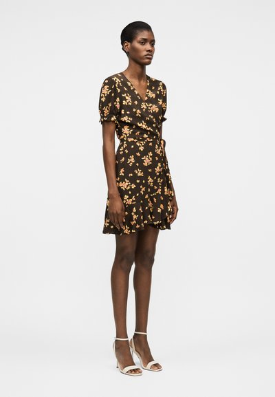 Γυναίκα που φοράει κοντό μαύρο φόρεμα τύπου wrap με κίτρινο λουλουδάτο μοτίβο, φουσκωτά μανίκια, βολάν στο τελείωμα και λευκά πέδιλα με λουράκι στον αστράγαλο και ψηλό τακούνι.