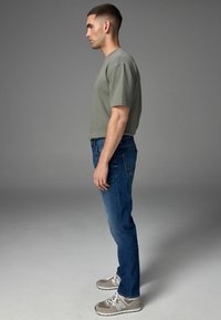 Khaki T-shirt met korte mouwen en een textuurfinish, gecombineerd met blauwe denim jeans. Accessoires zijn grijze sneakers met witte accenten.