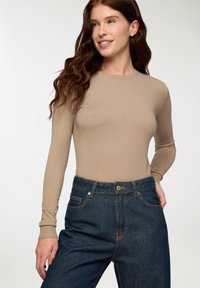 Maglia beige a maniche lunghe realizzata in un tessuto morbido, design aderente, abbinata a jeans di denim blu scuro a vita alta con cuciture a contrasto e tasche.
