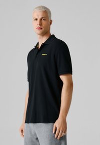 Mann mit kurzen blonden Haaren, der ein schwarzes Red Bull Racing Polohemd und graue Hosen trägt, stehend vor einem schlichten grauen Hintergrund.