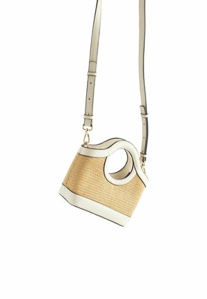 Kvinde i beige, strikket, ærmeløs top og hvid nederdel, der holder en lille, tan og hvid vævet håndtaske bag ryggen.