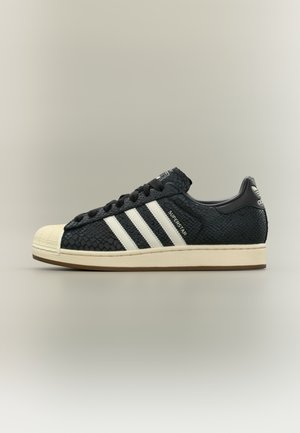 Black textured sneaker with white stripes, featuring a rubber shell toe and a flat lace design. The heel has branding accents.