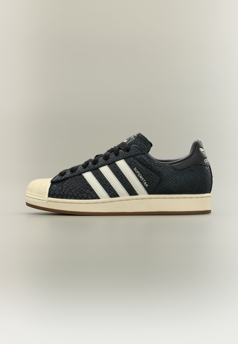 Μαύρο αθλητικό παπούτσι με υφή και λευκές ρίγες, με λαστιχένια προστασία στα δάχτυλα και επίπεδο σχέδιο κορδονιών. Η φτέρνα έχει λογότυπα.