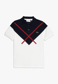 Polo tričko s bílým tělem, navy modrým vrškem a červenými diagonálními pruhy. Obsahuje klasický límeček a logo Lacoste na hrudi.