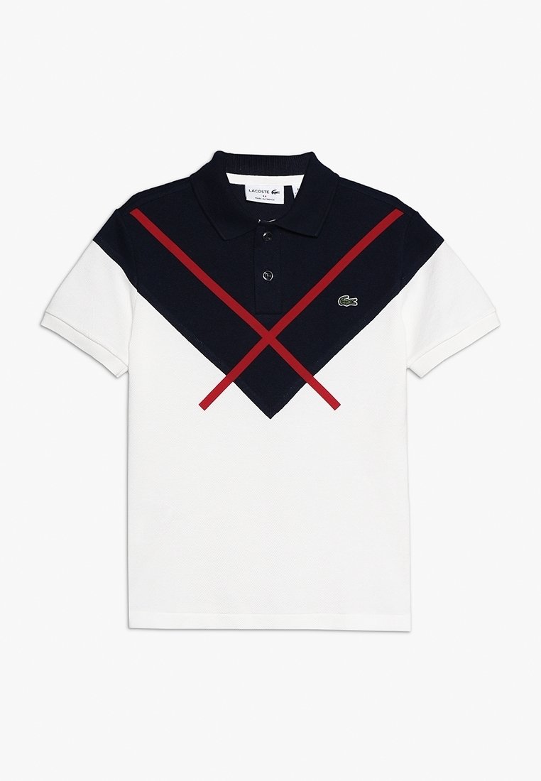 Polo tričko s bílým tělem, navy modrým vrškem a červenými diagonálními pruhy. Obsahuje klasický límeček a logo Lacoste na hrudi.