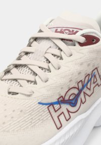 Sapatilha atlética bege com um cabedal texturizado, apresentando um logo "HOKA" em vermelho e uma linha de destaque azul. Sola branca e fecho de atacadores.