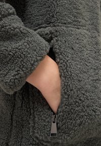 Chaqueta de forro polar gris con una superficie texturada y esponjosa, que cuenta con un bolsillo con cremallera, mostrando un primer plano del tejido y el detalle de la cremallera.