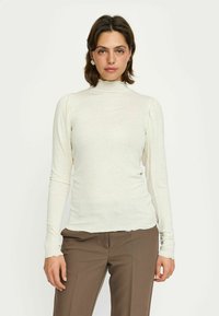 Top de culoare crem deschis, cu mânecă lungă și guler înalt, având umeri puțin bufanți și o textură moale, asortat cu pantaloni maro.