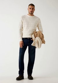 Hvid strikket sweater med tekstureret mønster, mørkeblå jeans, holder en beige jakke. Modellen har mørke sko på og står foran en lys baggrund.