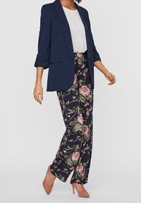 Marinblå blazer med strukturerad design, parat med blommönstrade byxor i mörka toner, framhämtade av rosa och gröna detaljer. Rosa klackskor fulländar looken.