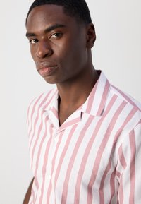 Camicia leggera a maniche corte con strisce alternate rosa e bianche. Presenta un colletto da campeggio e bottoni sul davanti. Texture del tessuto liscia.