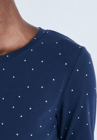 Närbild av en persons överkropp som bär en marinblå skjorta med små vita prickar och rund halsringning.
