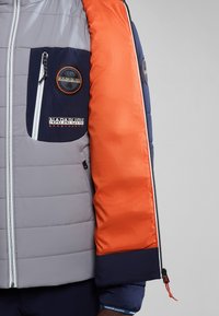 Giubbotto imbottito con corpo grigio trapuntato, maniche arancioni e dettagli blu navale. Presenta una tasca sul petto con zip e toppe con il marchio.