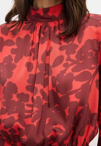 Rød bluse i stof med høj krave, læg og blomstermønster i mørkere nuancer af rød. Glat tekstur med et draperet design.