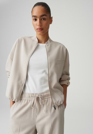 Femme aux cheveux tirés en arrière, portant une veste zippée beige et un pantalon assorti à cordon de serrage, avec un haut blanc côtelé, les mains dans les poches.