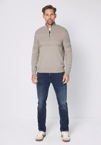 Beżowy sweter z dzianiny z półzamkniętym kołnierzem i poziomym wzorem w paski, połączony z ciemnoniebieskimi dżinsami i białymi trampkami.