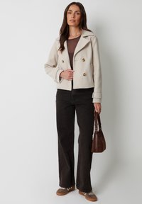 Beige cropped peacoat med dobbeltrækker i guld, blød tekstur, parret med mørke flared jeans og en brun håndtaske.