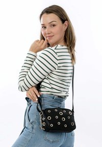 Borsa a tracolla nera con dettagli in occhielli metallici, forma rettangolare, materiale in tessuto e tracolla removibile, indossata sopra una camicia a righe.