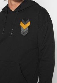Felpa nera con tasca frontale, caratterizzata da tre toppe in tessuto a forma di chevron nei colori grigio scuro, grigio chiaro e arancione, con chiusura a coulisse.