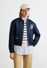 Jeune femme portant une veste bomber bleu marine sur un pull rayé et une chemise à boutons, une casquette beige et un pantalon kaki, debout avec une main dans la poche.