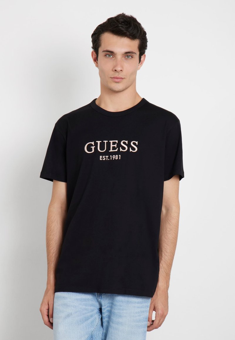 T-shirt noir en coton avec "GUESS" et "EST.1981" brodés en couleur crème. Encolure ronde, manches courtes, coupe décontractée.