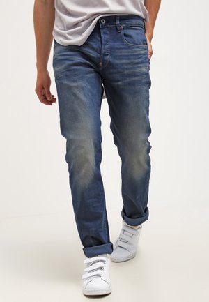 Homme portant un jean bleu délavé avec les revers roulés et des baskets blanches à velcro, marchant avec une main dans la poche.