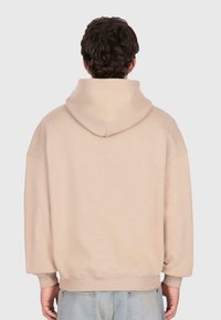 Un homme portant un sweat à capuche beige et un jean bleu clair, se tenant de dos contre un fond clair uni.