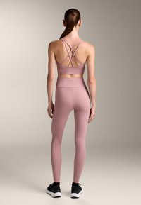 Súprava športového podprsenky a legín v svetloružovej farbe, s dizajnom krížových popruhov a hladkou textúrou, doplnená čiernymi športovými topánkami.