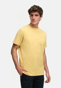 Gul bomulls t-skjorte med rund hals, korte ermer og en liten hvit logo på forsiden, paret med mørke bukser.