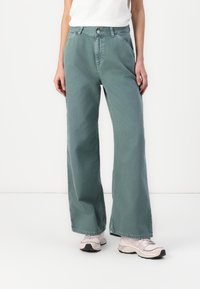 Videbenbukser i en blød teal-farve, lavet af denim med en glat tekstur. Har en knaplukning og sidelommer. Båret med lyse sneakers.